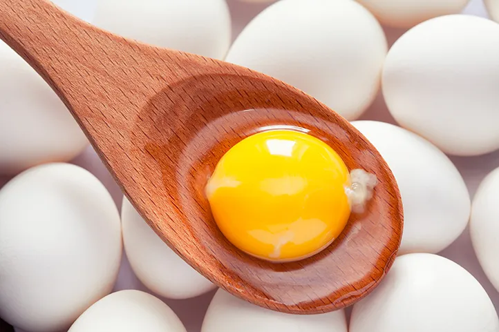 an egg yolk sitting in a wooden ladle suspended over a group of white eggs
